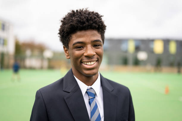 Head and shoulder front view of 15 year old student in shirt, necktie, and navy blue blazer standing outdoors on sports field and smiling at camera.